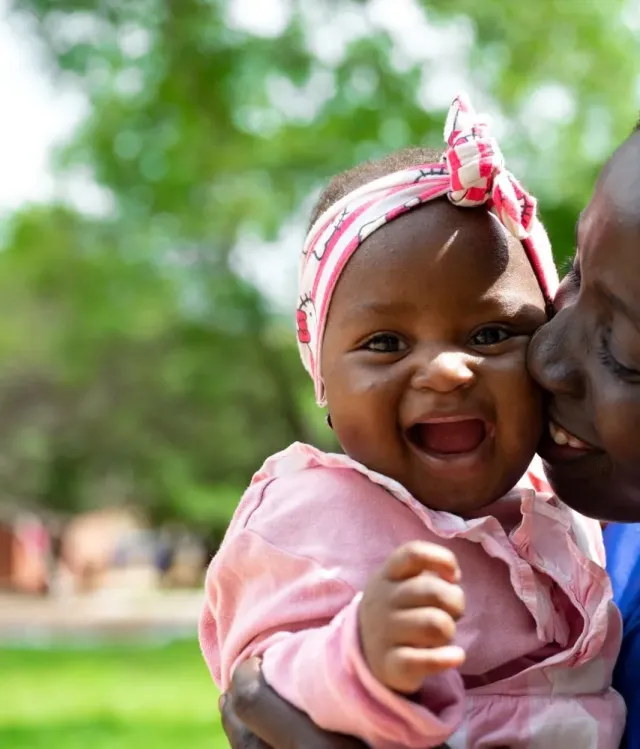 Smiling mother in a blue shirt kisses her baby daughter’s cheek in a park; the baby wears pink and a bow in this joyful, loving scene.