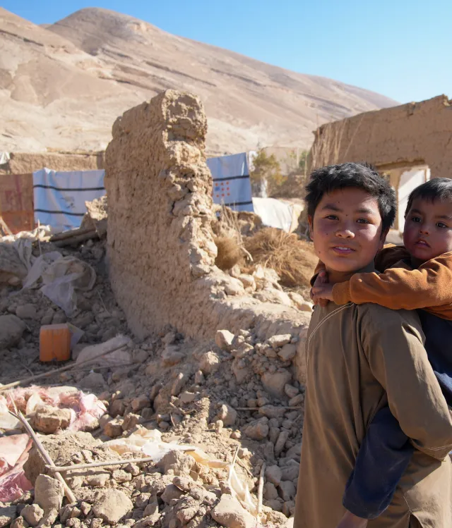 Boy carrying his younger brother on his back beside earthquake rubble in Afghanistan.