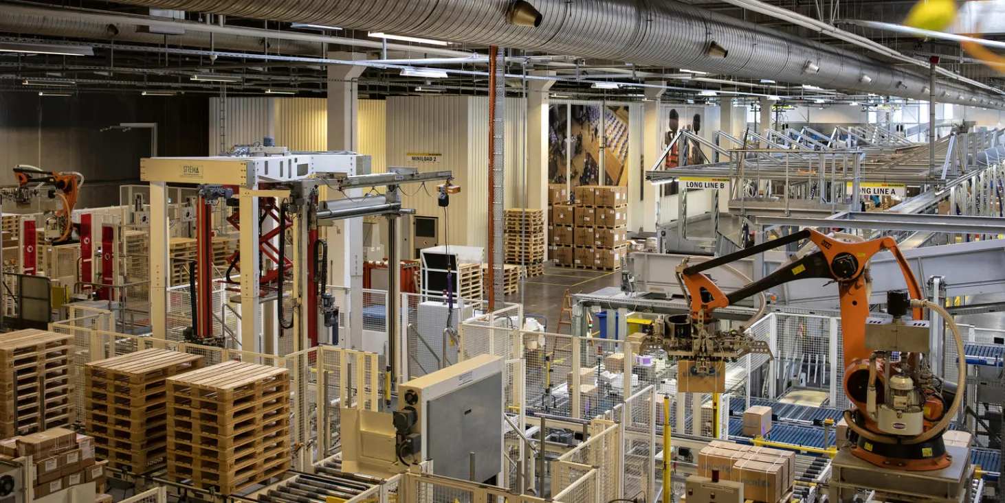 Image of warehouse with skids piled about 5-feet high, and an orange crane is moving back and forth in the warehouse. 