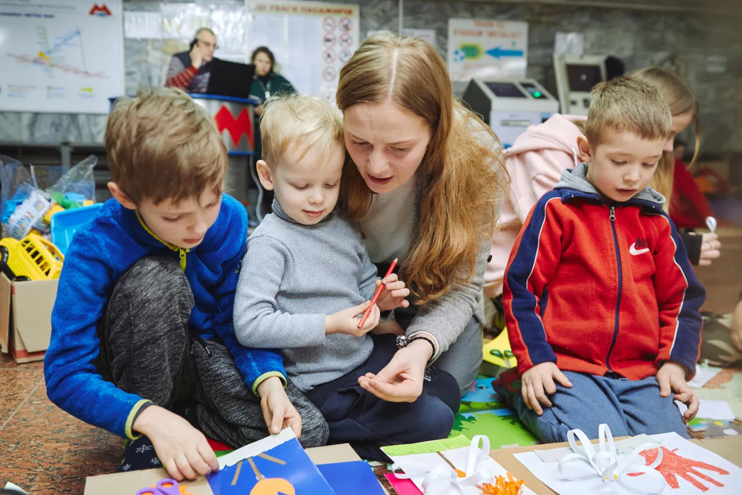 Ukrainian volunteer Anastasiia, with long blonde hair and grey shirt, sits with three Ukrainian boys in a UNICEF support centre. They are all seated, doing arts and crafts. The first boy has dirty blonde hair, with blue shirt and yellow accents, and grey jeans or sweatpants. The second boy has light blonde hair, with a light grey shirt and blue jeans. The third boy has a red Nike sweater with a blue stripe on the sleeves. Another blonde woman is behind the boy in red, wearing a pink sweater. 