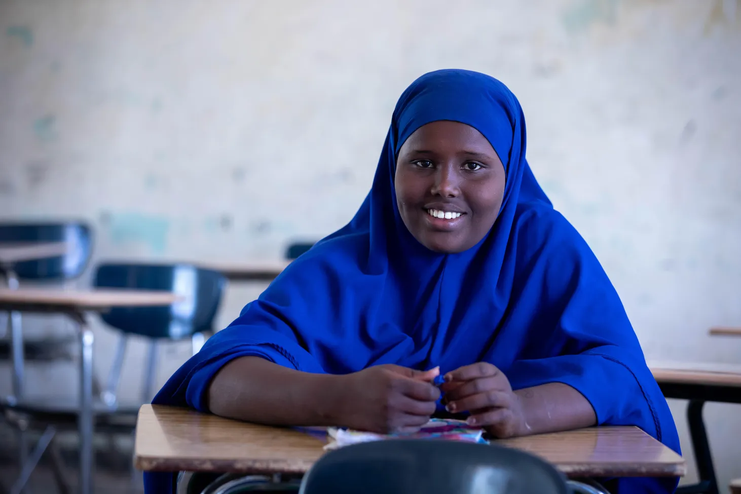 A girl wearing a blue headscarf sits on a desk in a classroom and looks straight at the camera. She is smiling, a book is lying open in front of her. 