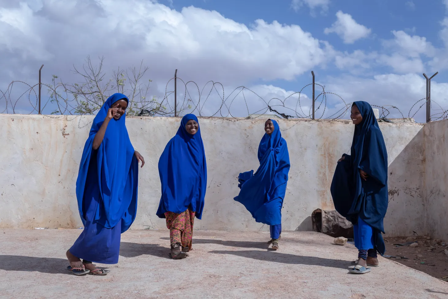 Four girls in blue stand in a school courtyard, all are smiling or laughing. 