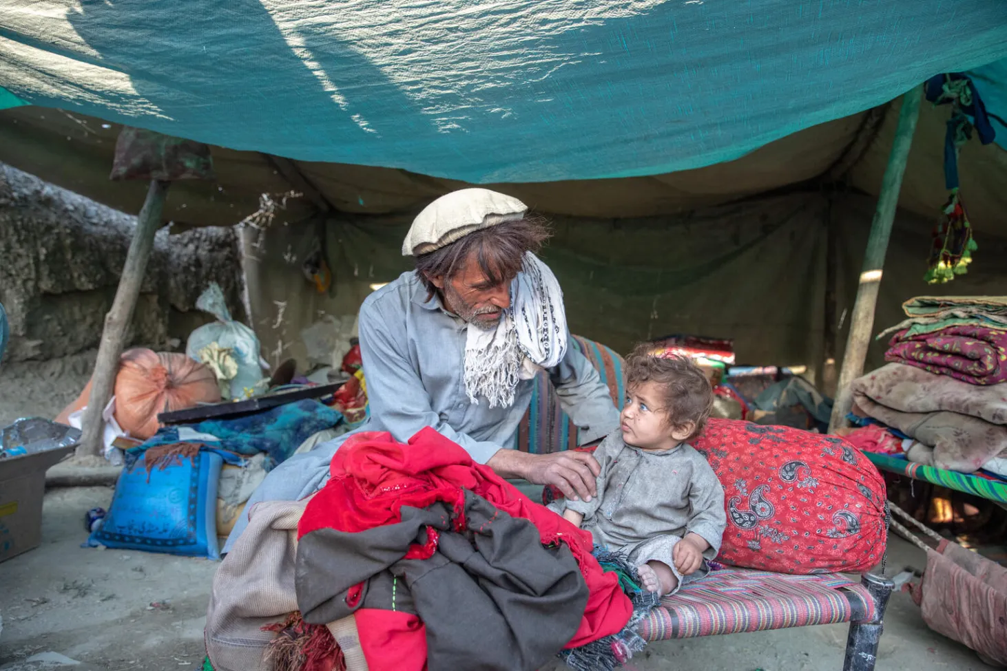 A man sits on a woven bed with an infant next to him. They are looking at each other, behind them is a tent, like a makeshift shelter, with belongings lying in sacks and piles. 