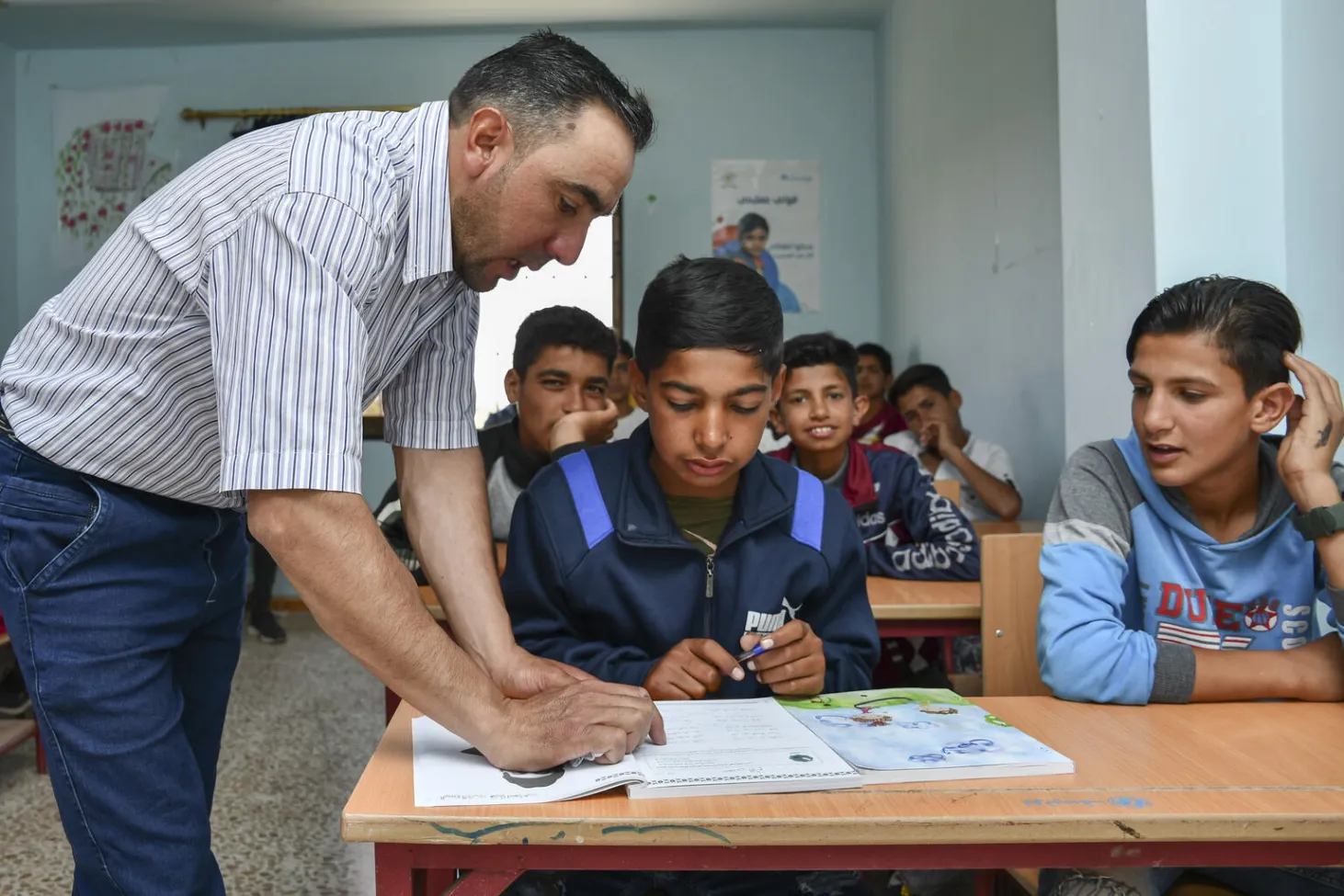 A school teacher leans over the desk of two students who have a book open in front of them.