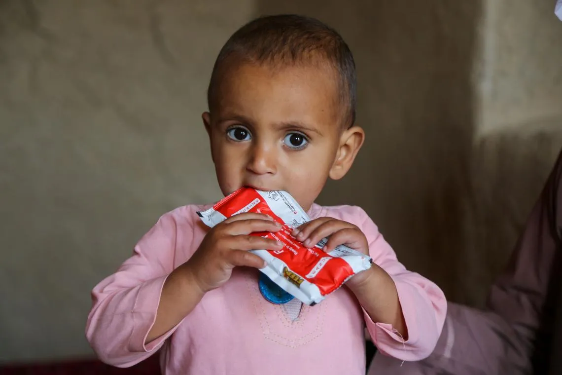 A toddler in pink clothing is holding a red packet of ready-to-eat-therapeutic food. The photo is zoomed in, we can see the toddler from the chest up. He is looking at the camera.  