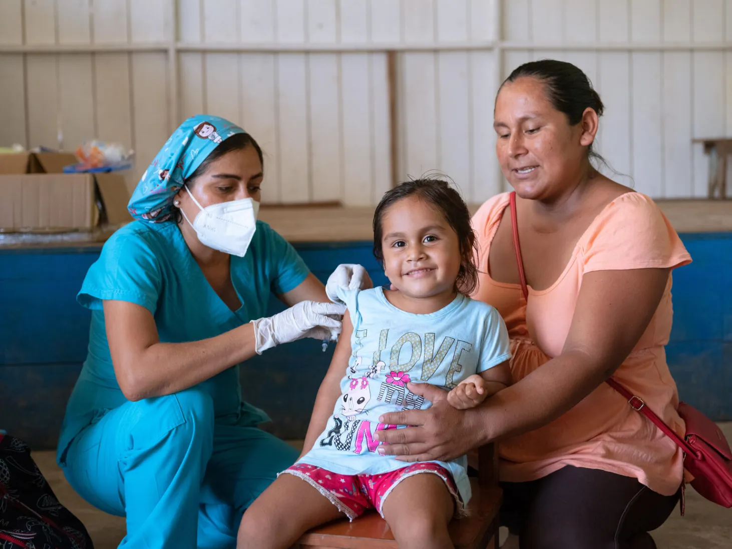 A three year old girl in shorts and a tshirt sits in between her mother and a nurse. The nurse is administrating an injection to the girls arm. The mother is holding the girl. The girl is smiling and looking straight at the camera. The setting looks like a make-shift mobile health clinic. 