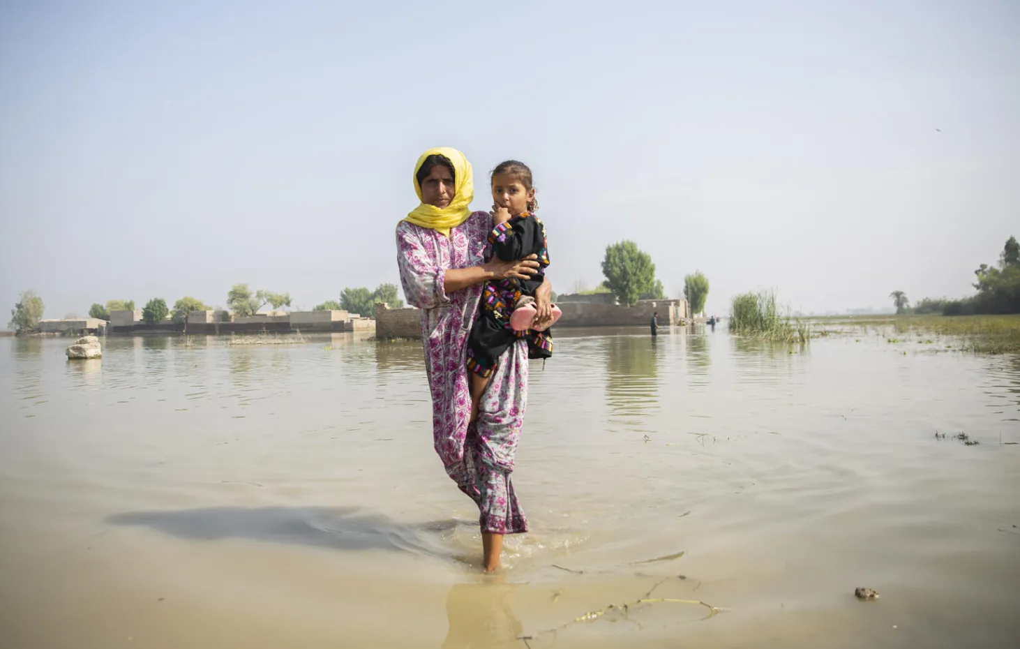 A woman walks though ankle-deep water holding a child in her arms, behind her submerged houses can be seen with water covering the entire landscape. 