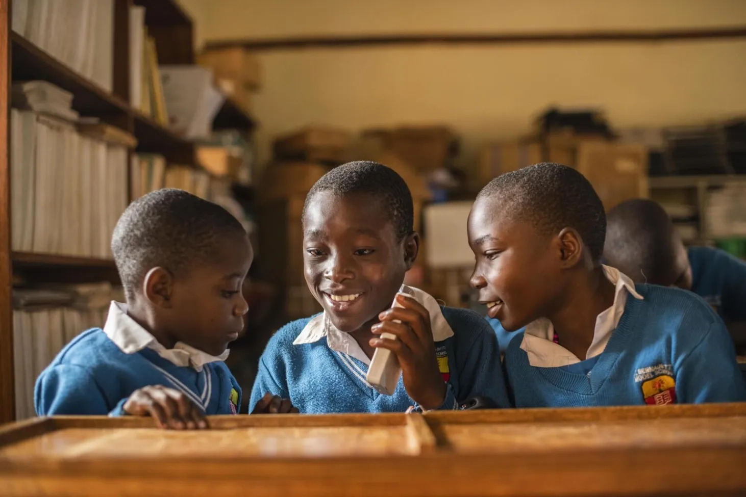 Three boys in blue school uniform sweaters sit at a desk, the boy in the middle is holding a listening device up to his ear and smiling while the two boys next to him are turned towards him. 