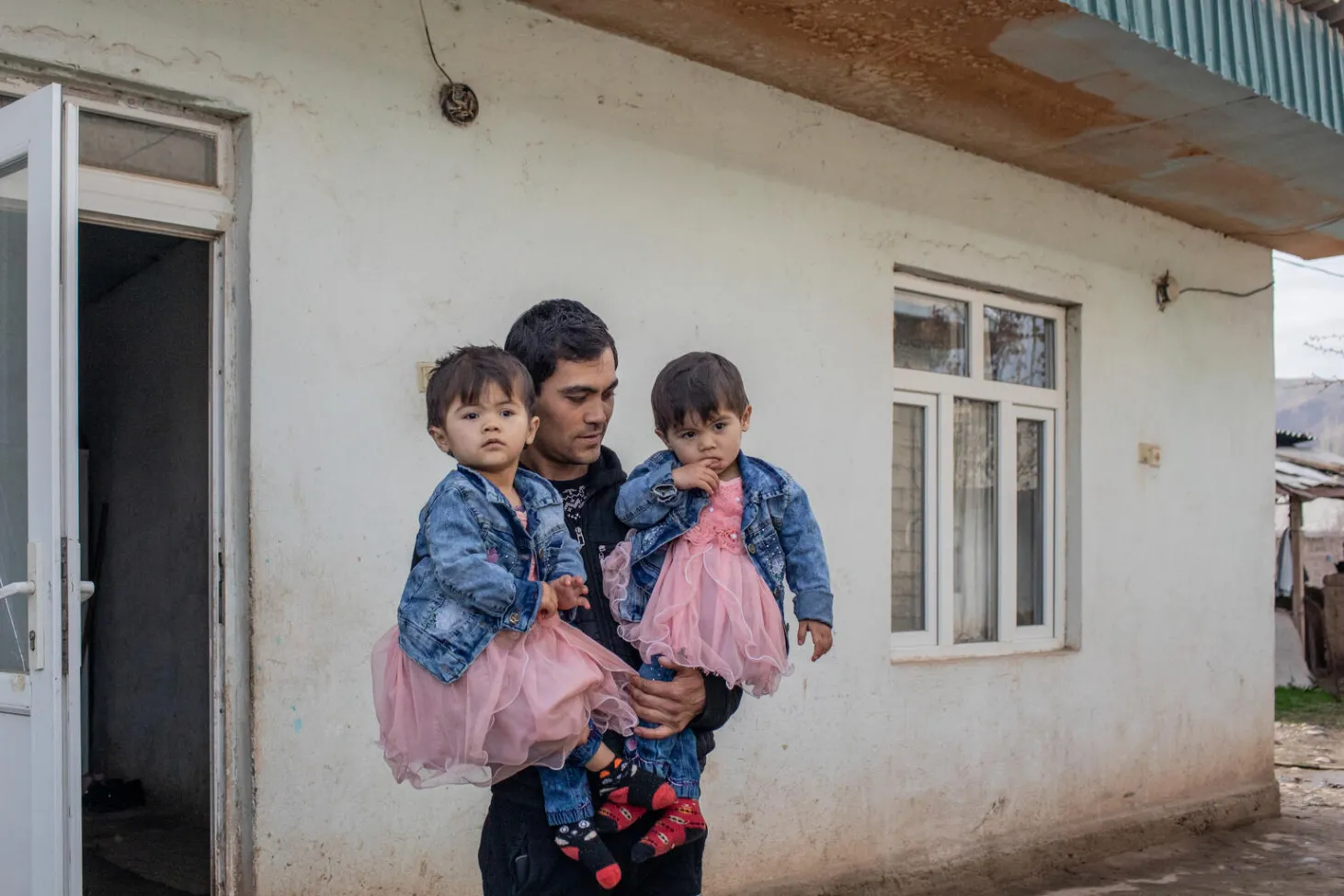 Papa Namazqul tient ses deux filles dans ses bras à l’extérieur de leur maison. 