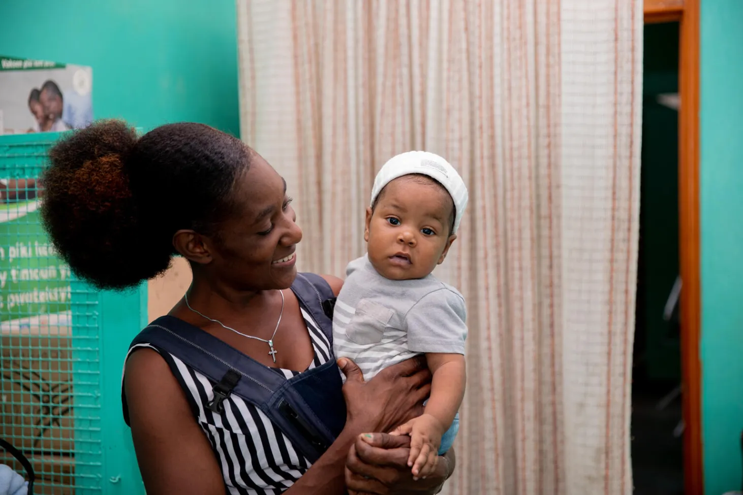 A woman holds an infant and is smiling. The infant is looking at the camera while the woman is looking at the child. They are in a clinic-looking setting. 