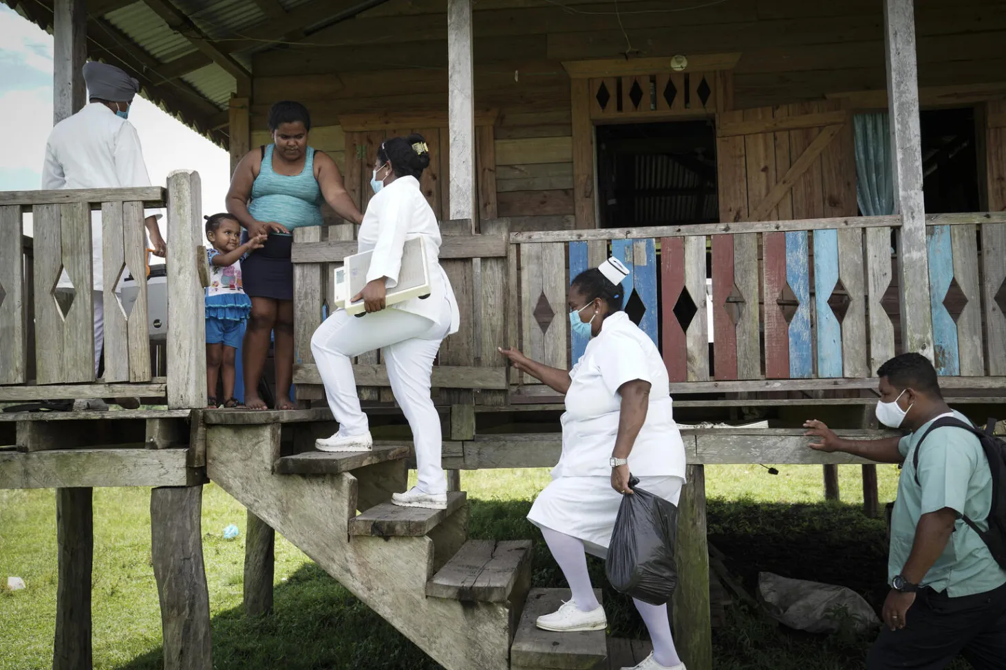 Three women, two in white nurses uniforms, climb up stairs to the terrace of a home, where a child and women are waiting for them. 