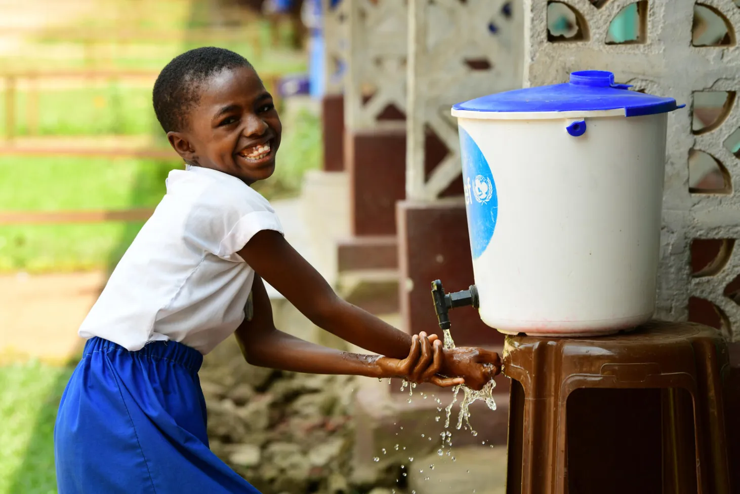 a child washer her hands at a portable handwashing station - a bucket with a tap - she is smiling and looking at the camera. 