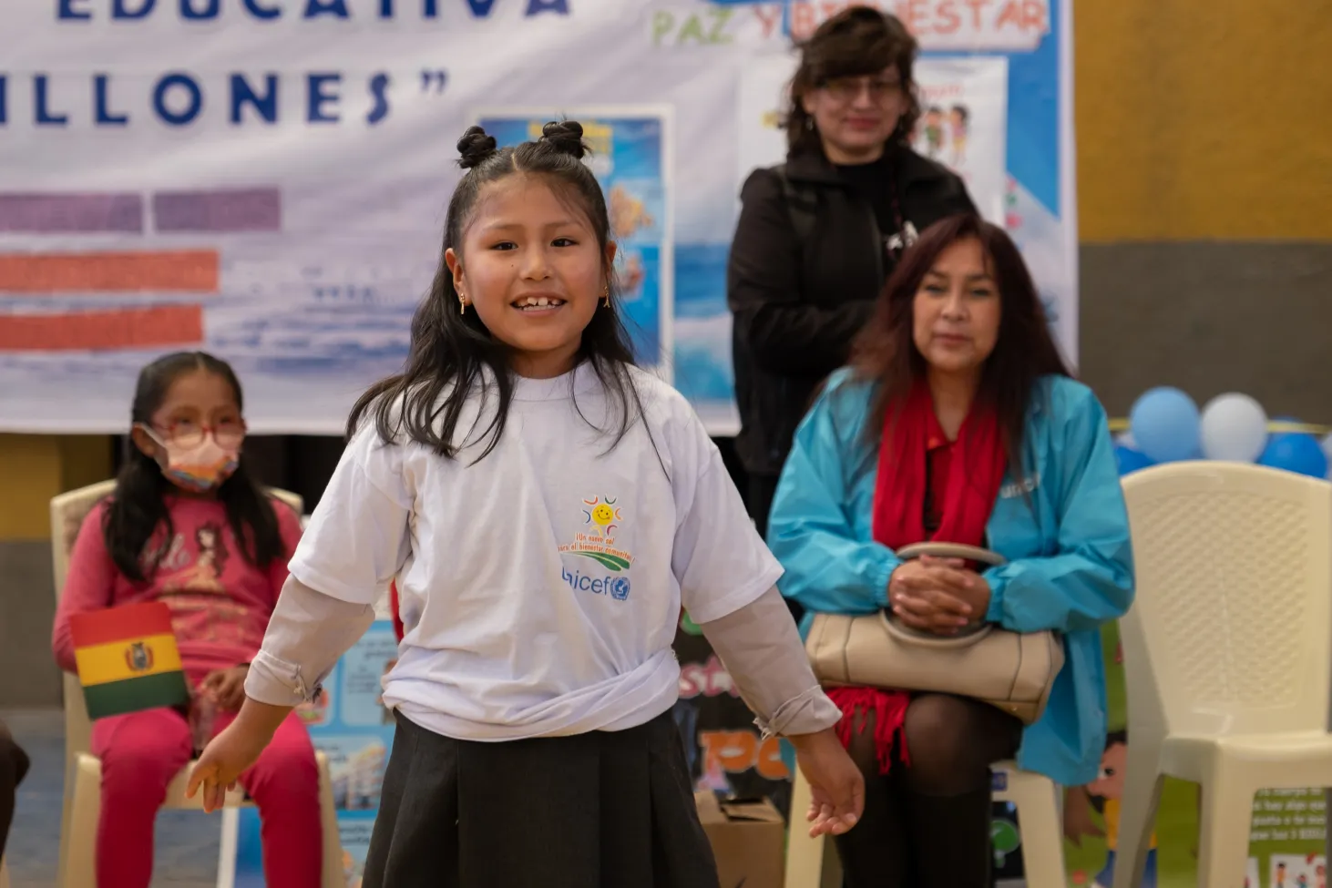 A young girl in an auditorium at a school rally in Bolivia. She is looking at the camera and smiling.  