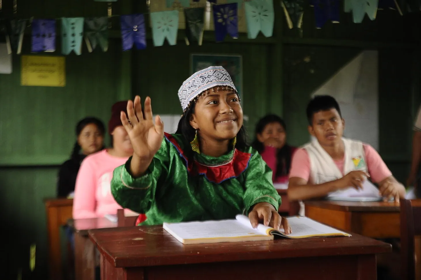 A girl sits at a desk in a classroom with others behind her. She is raising her hand and smiling while wearing traditional Peruvian headband.  