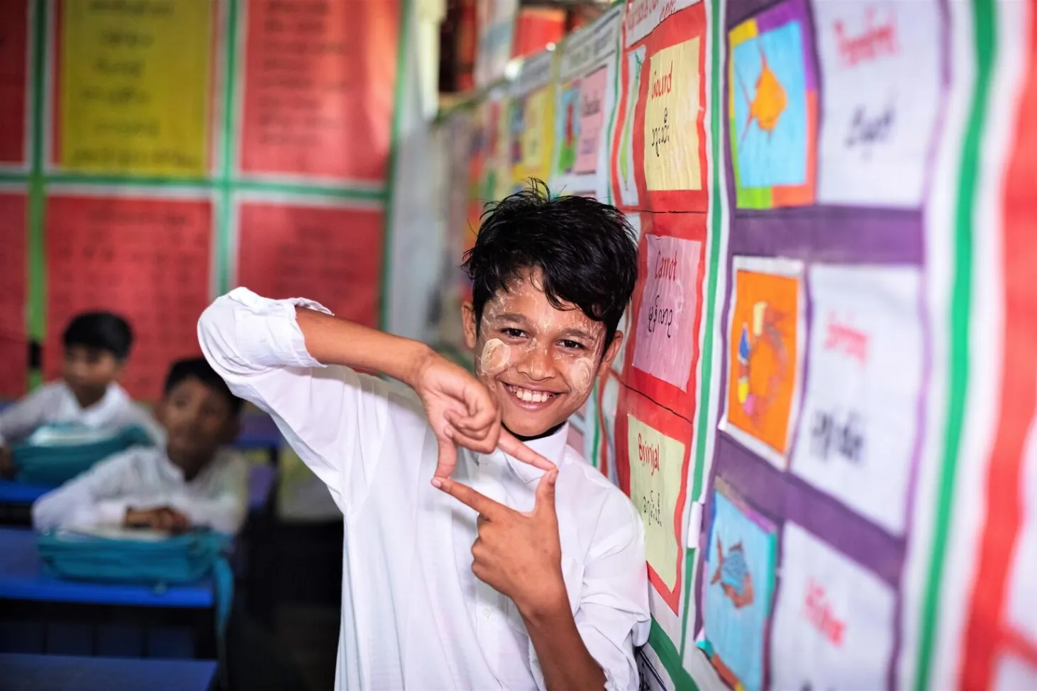 A boy uses his hands to create a square photo sign. He is in a classroom setting standing next to bulletin board with many colourful sheets pinned on it. He is wearing a white uniform shirt. In Cox's Bazaar, Bangladesh. 