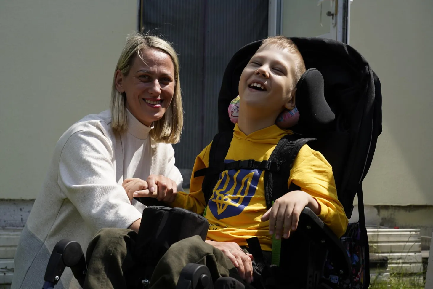 A mother leans next to her son in a wheelchair, both are smiling and looking at the camera. In Ukraine. 