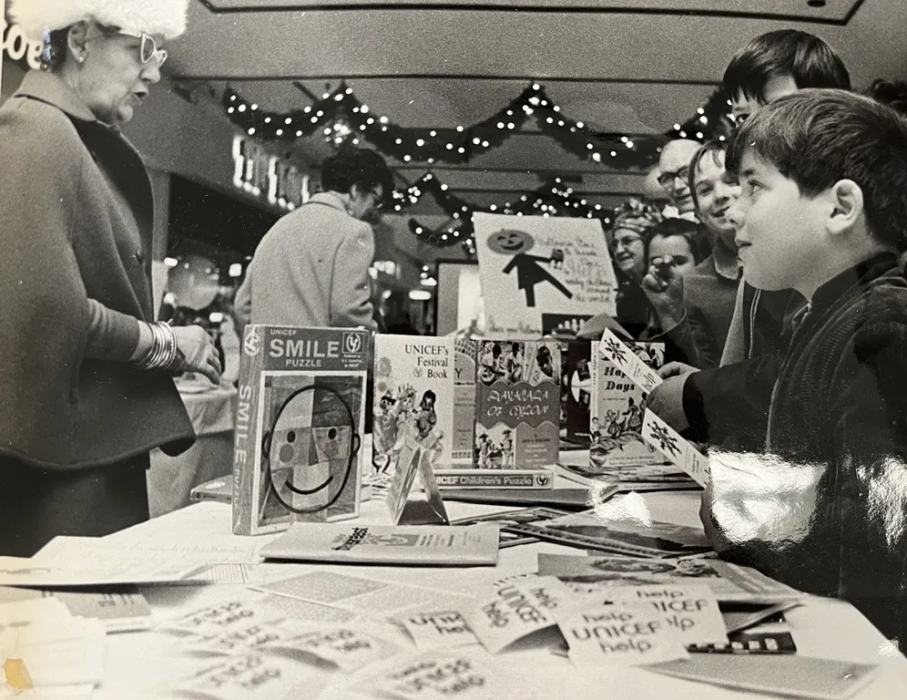 Photo en noir et blanc de deux enfants regardant une femme derrière une table qui vend des articles de l’UNICEF.