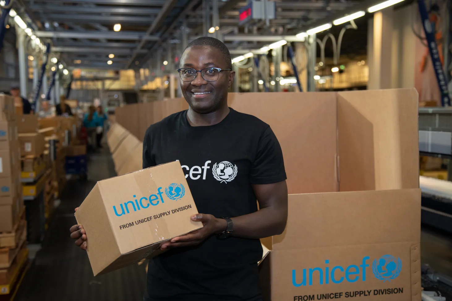 A man holds a cardboard box that says UNICEF on it, behind him more boxes are stacked. 