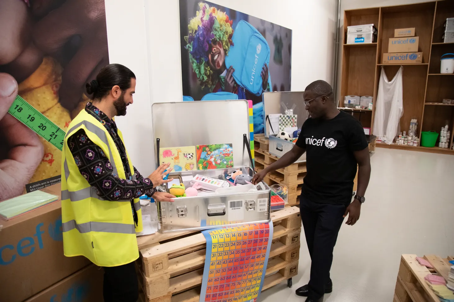Two men stand besides a metal box that is open and full of school supplies. 