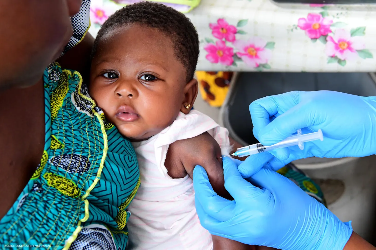 Une petite fille regarde l’objectif alors qu’elle reçoit un vaccin dans le bras administré par une personne portant des gants bleus. 