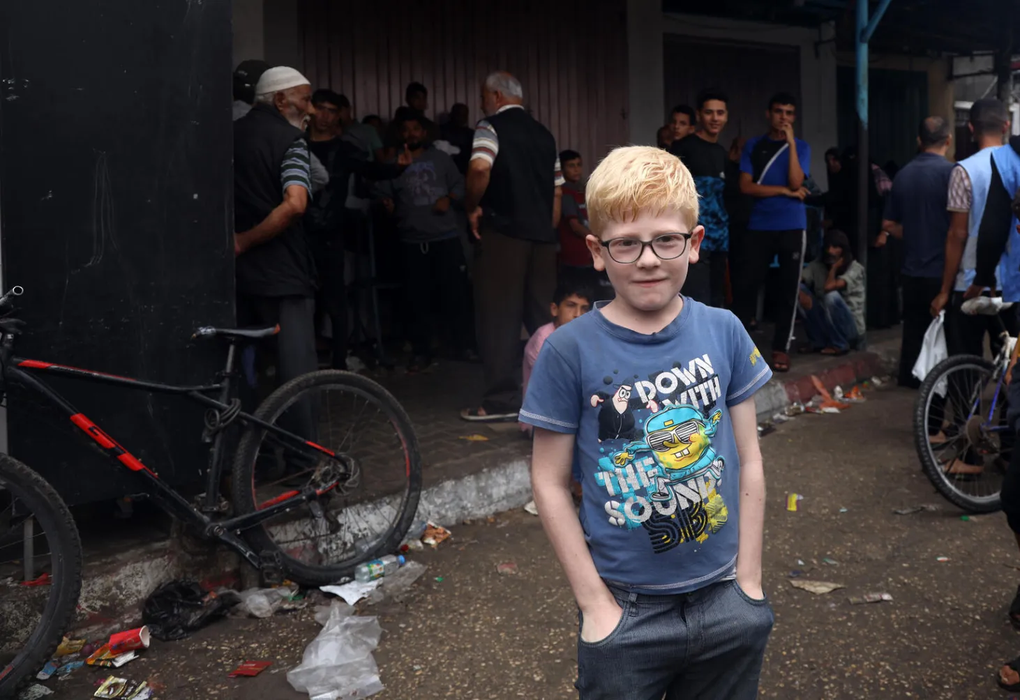 Un enfant, les mains dans les poches, regarde l’objectif. Derrière lui, des hommes en file attendent pour se procurer du pain.