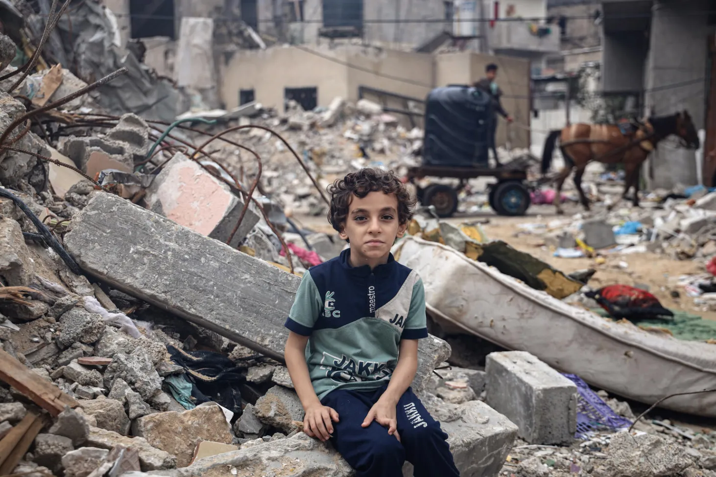 Un enfant assis sur les décombres de la maison familiale regarde l’objectif. On voit derrière lui tous les débris et gravats.