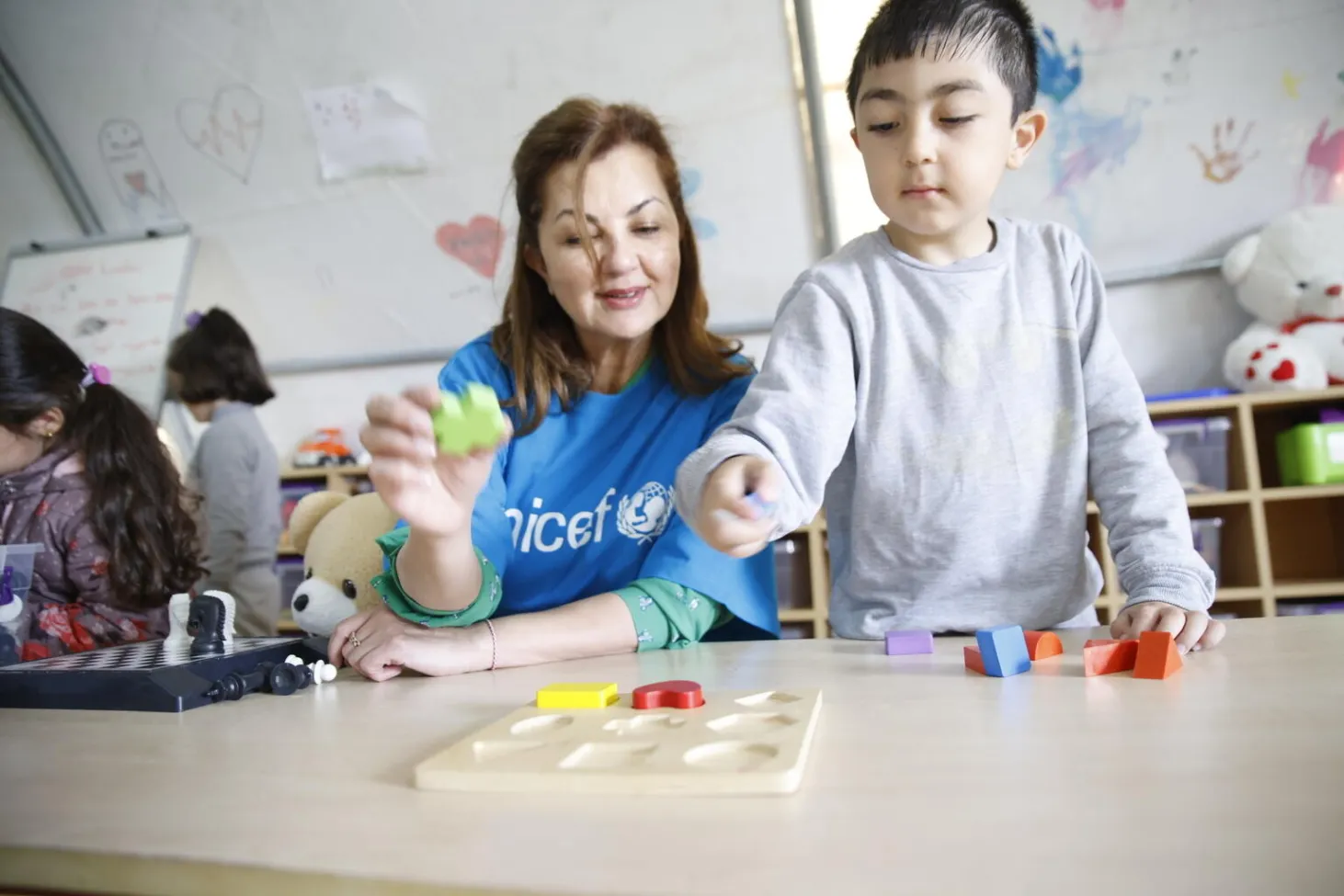 A woman sits at a table with a child who is standing up and showing some toys to her.