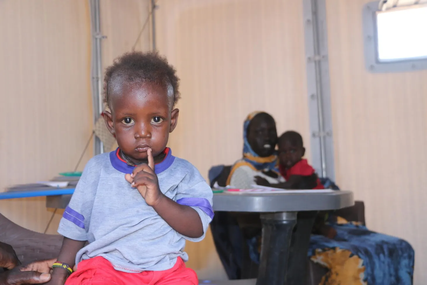 A child looks directly at the camera, one finger on his lips. He looks a little tired.