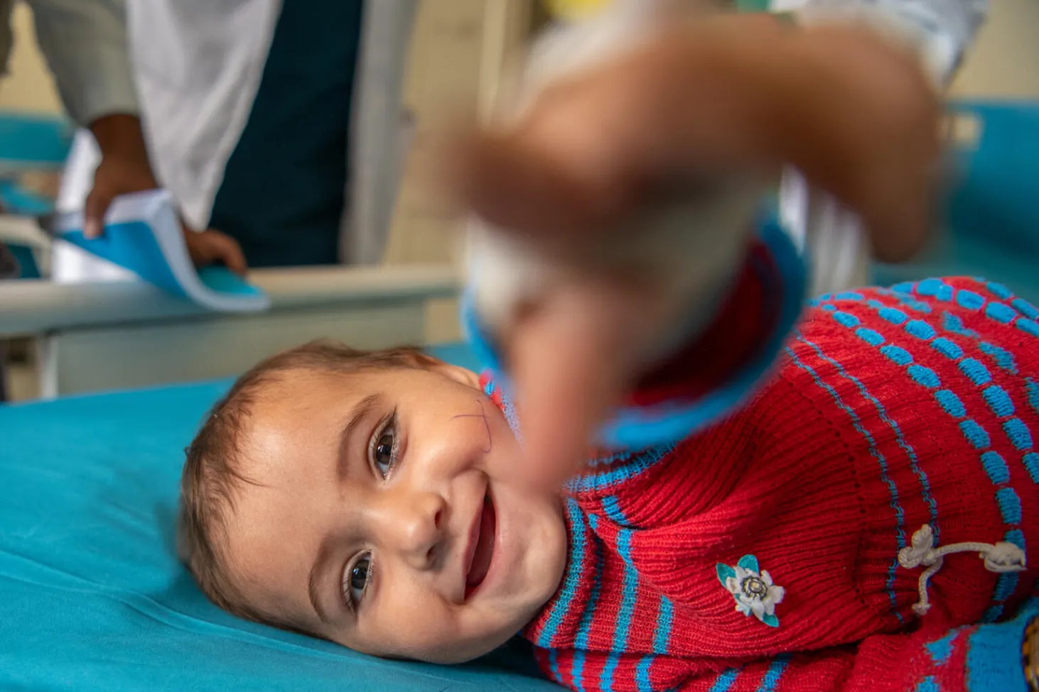 An infant on a bed extends his arm out towards the camera while he is lying down. He is smiling.  