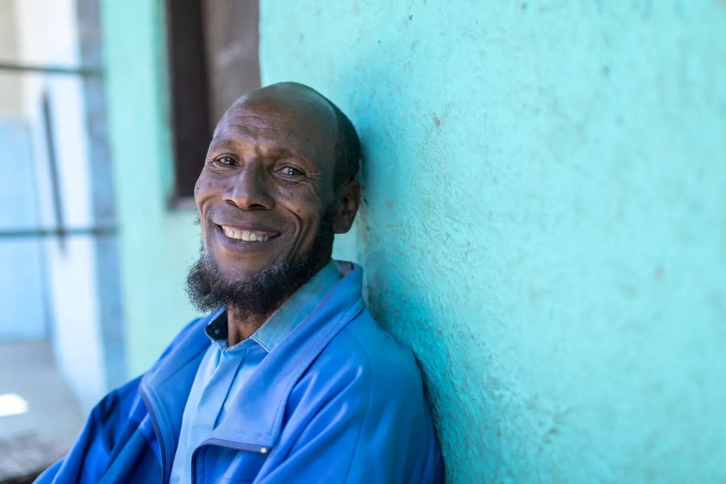 Azia's father looks at the camera smiling, he is leaning against a green wall. 