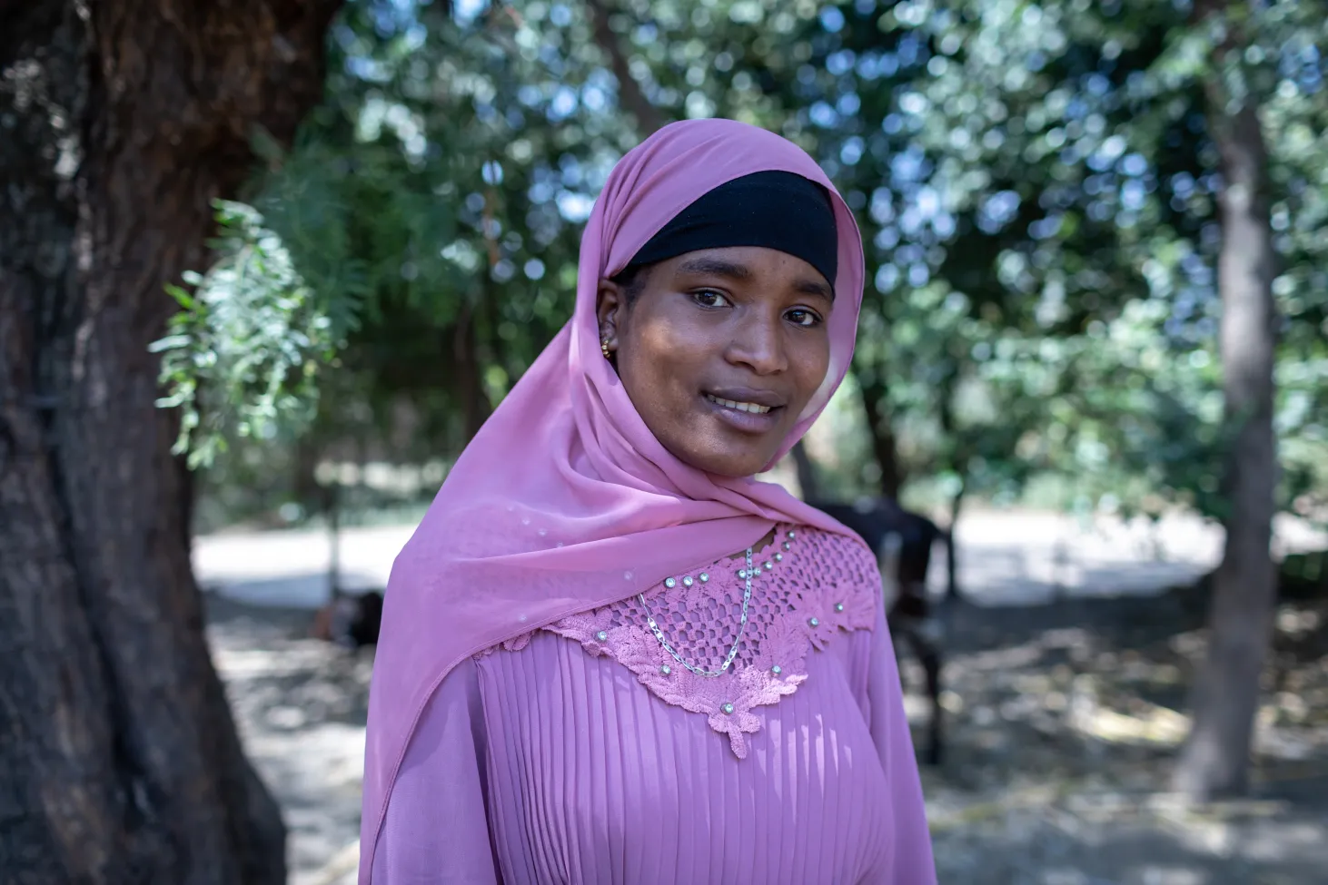 An adolescent girl in pink clothes looks directly at the camera with a slight smile. She is in an outdoor setting.