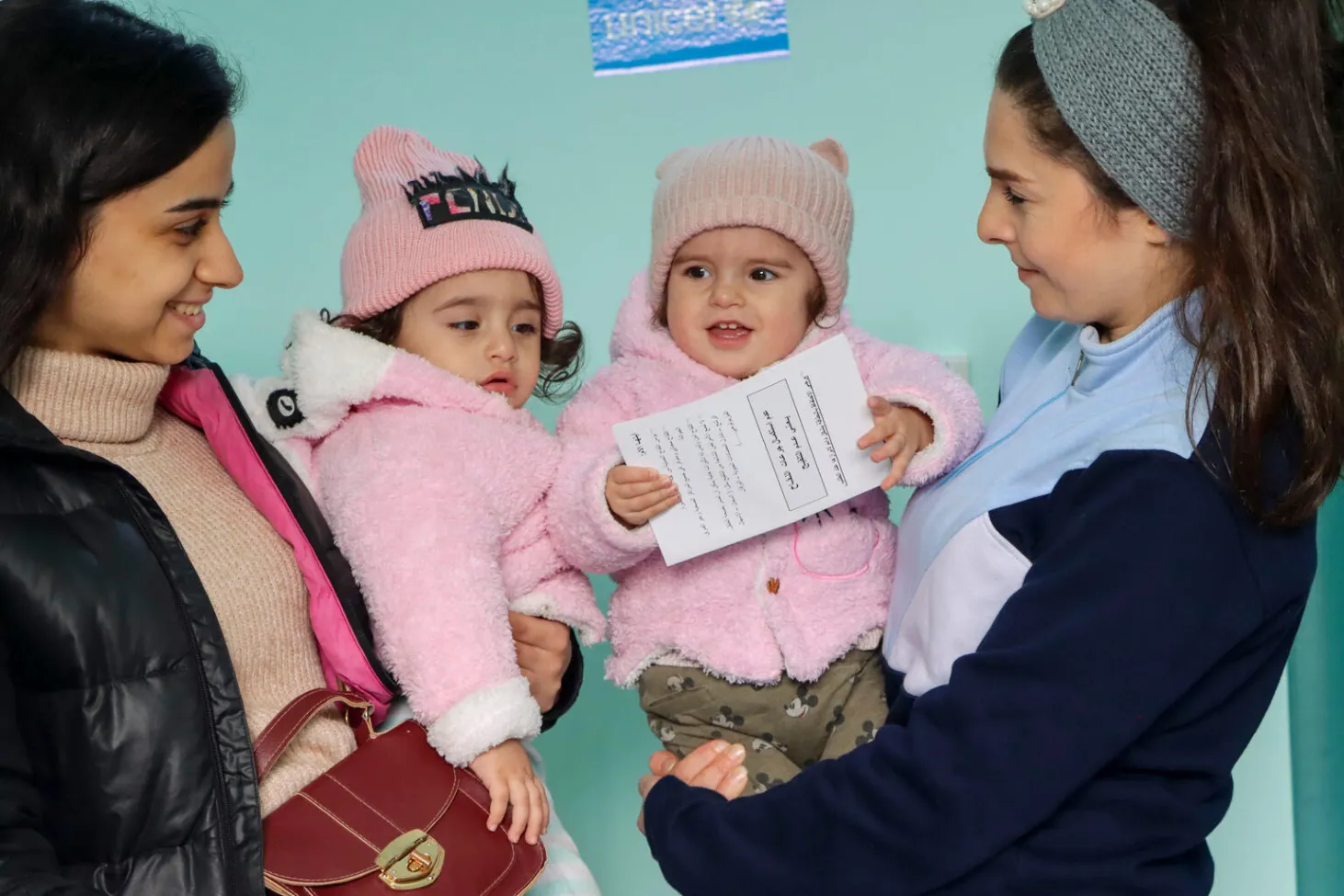 Des sœurs jumelles âgées d’un an dans les bras de leur mère et de son amie dans la salle de vaccination.