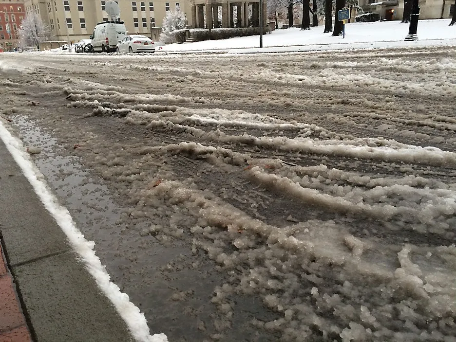 A slushy street covered in dirty snow in a city with buildings and parked cars in the background.