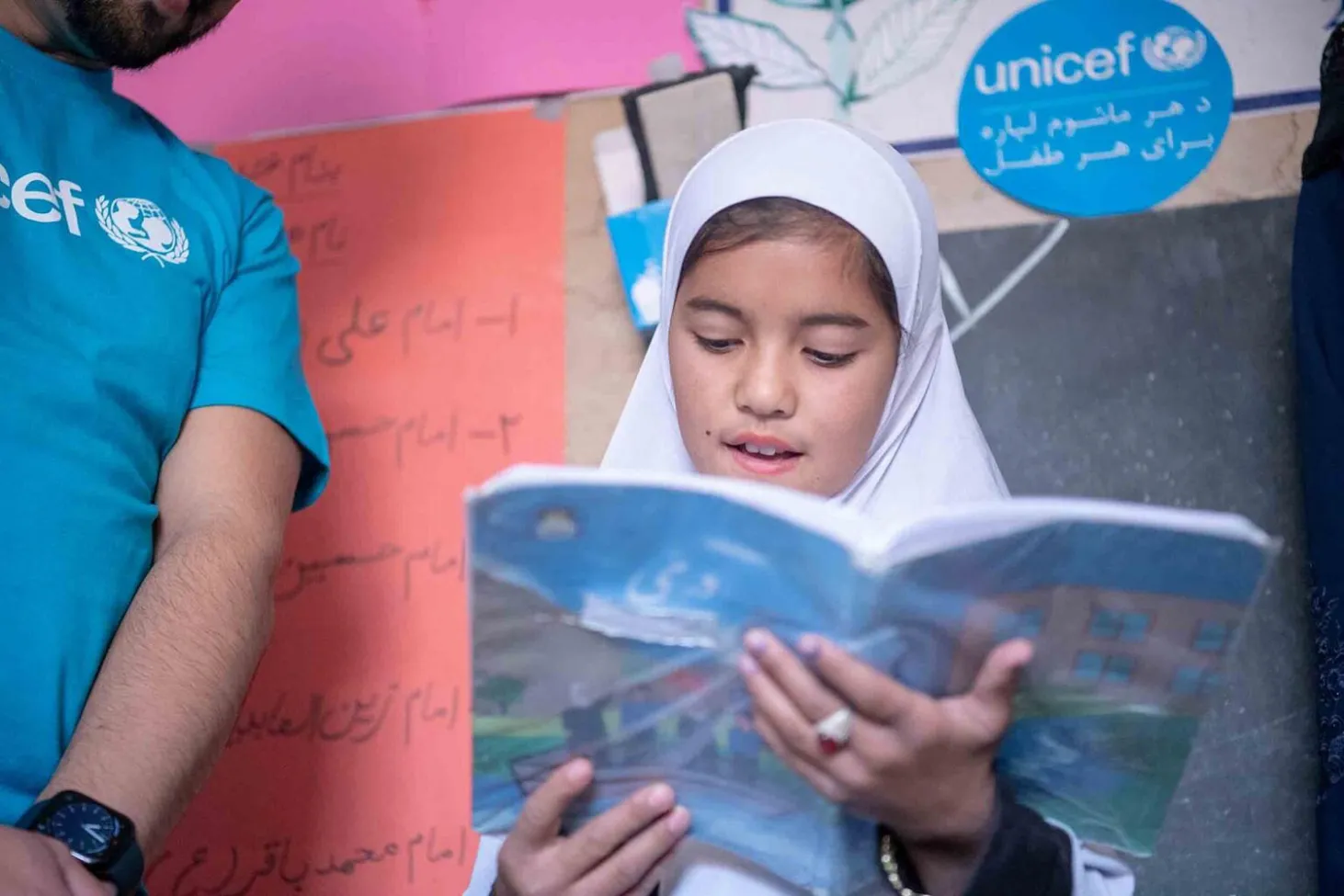 In a classroom, a girl wearing a white headscarf reads from a book as a UNICEF worker stands beside her.
