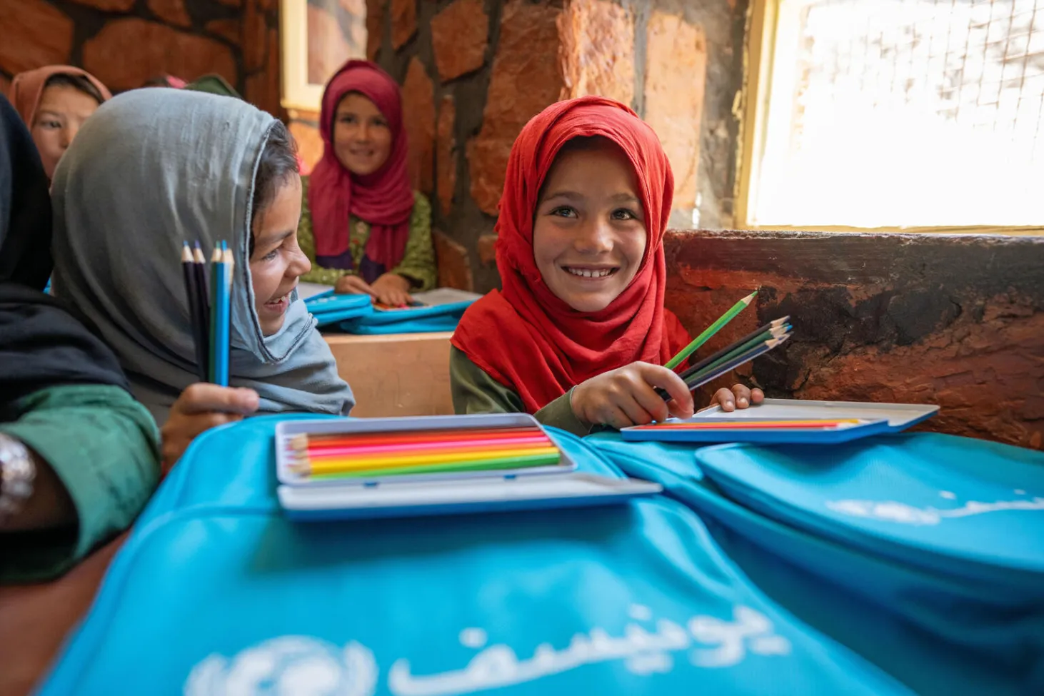 In a classroom, a group of girls smile while sitting at their desks that have new blue UNICEF backpacks and sets of pencil crayons on them. 