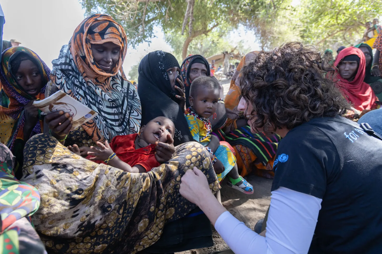 A group of women in colorful clothes and headscarves sit outdoors under trees, one holding a baby. A person in a UNICEF tshirt interacts warmly with them.