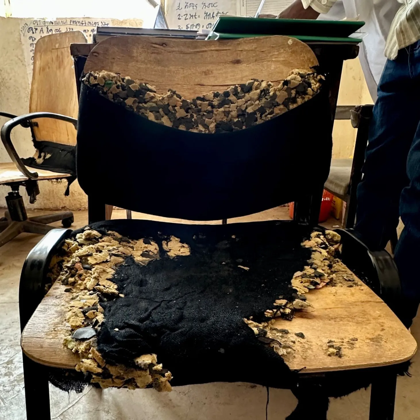 A worn-out chair with torn black fabric and exposed foam sits in a dimly lit room. Nearby, a person’s figure is partially visible.