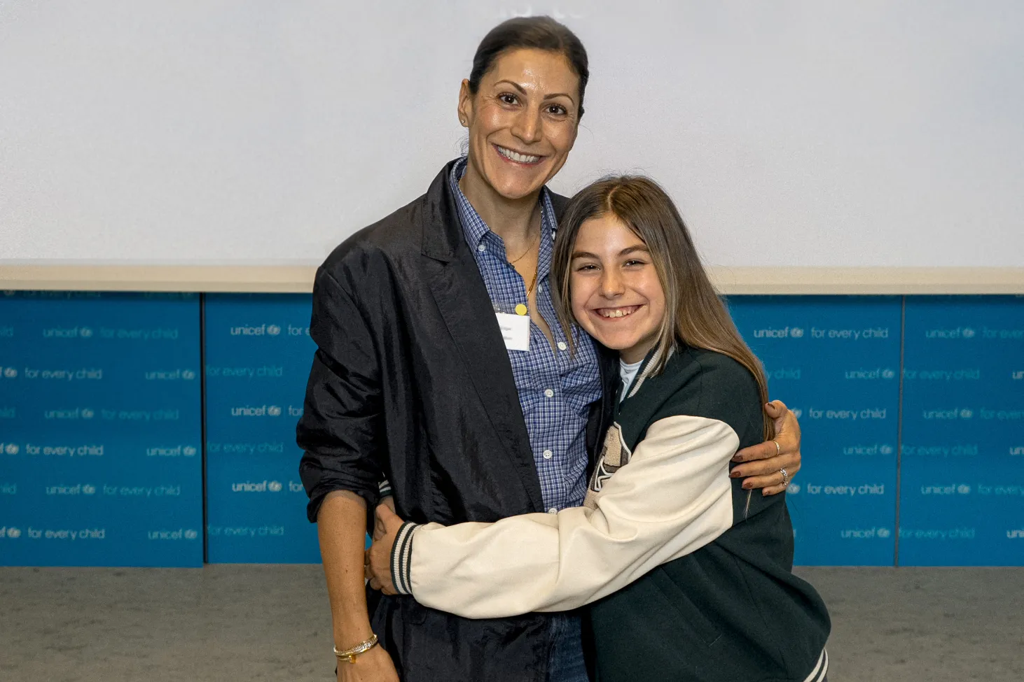 A woman and a young girl smile broadly, the girl and woman have their arms around each other by side in front of a backdrop displaying UNICEF's logo. The setting conveys a positive and friendly atmosphere.