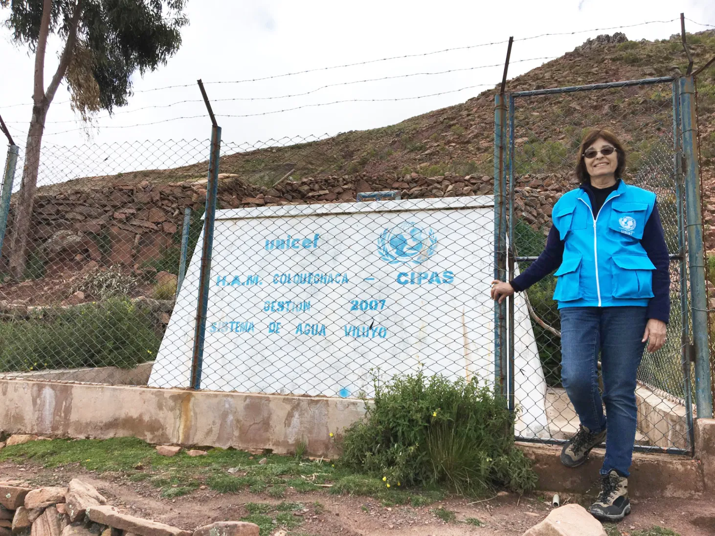 Susana wearing a bright blue UNICEF vest stands beside a chain-link fence in front of a sign with text and a logo, set against a rocky hillside.