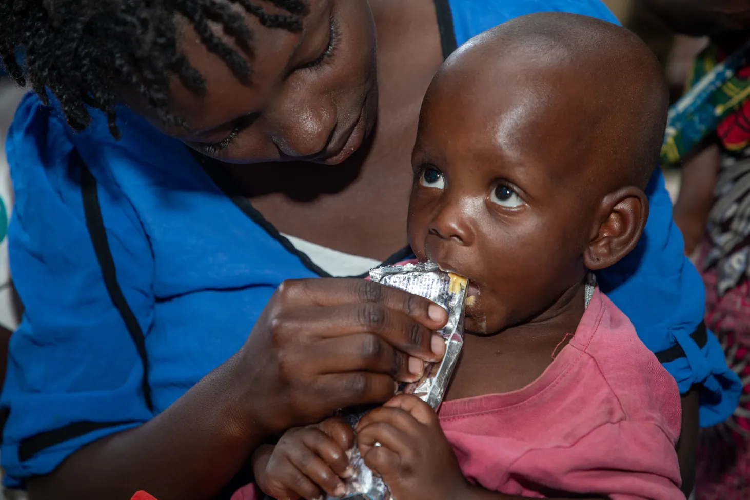 Close-up of a young child holding and eating from a small packet of RUTF with the help of his mother