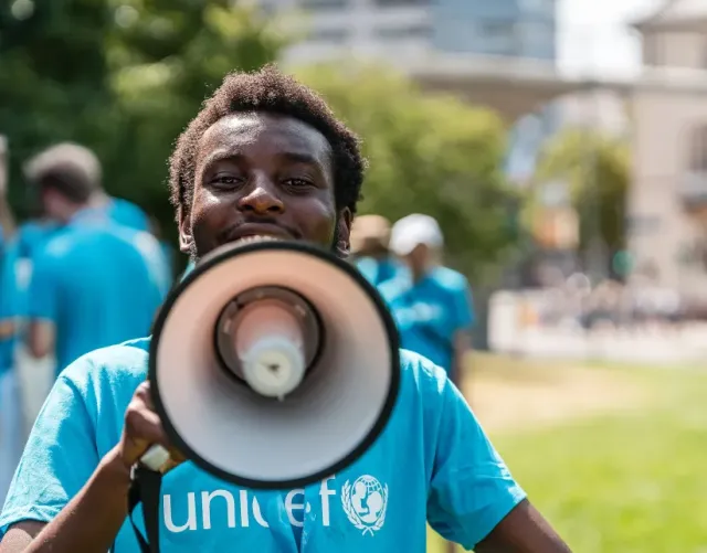 Smiling UNICEF volunteer in a blue shirt speaks through a megaphone outdoors, with a community gathering, lawn, trees, and city buildings behind.