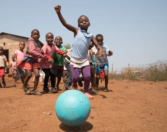 Children in a rural village run joyfully toward a turquoise soccer ball on dry red earth, with a simple brick house and trees behind them.