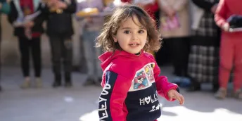 Young girl in a pink tracksuit smiling and running outdoors, with people gathered behind her.