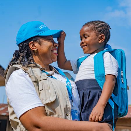A smiling UNICEF worker stands outside in the sunshine holding a child who is smiling and reaching for the worker’s UNICEF hat.
