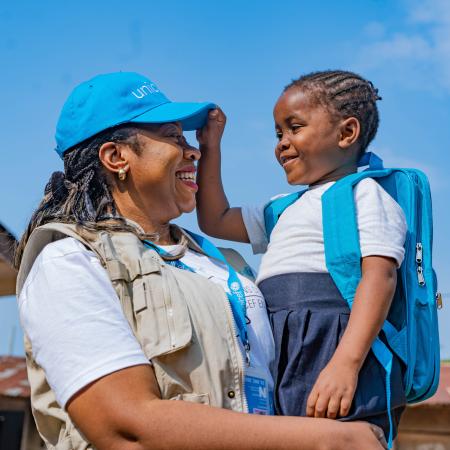 A smiling UNICEF worker stands outside in the sunshine holding a child who is smiling and reaching for the worker’s UNICEF hat.