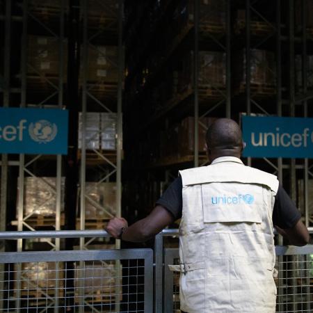 A man stands at a railing with his back to the camera, overlooking a warehouse staked with boxes. He is wearing a vest that says UNICEF.