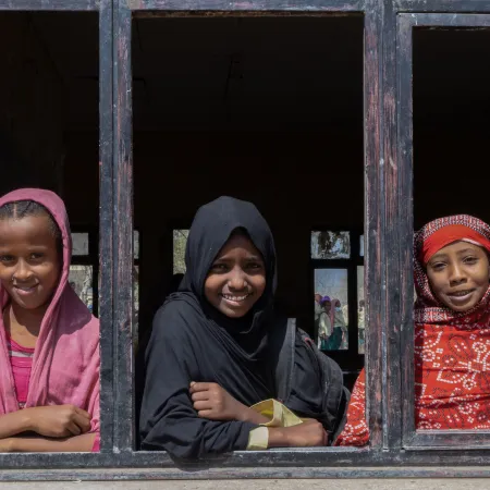 Three young girls with colourful head scarfs on, look out of a wooden window frame - all are smiling.  