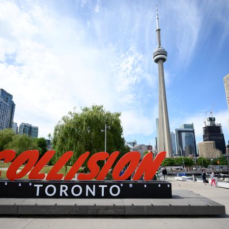 A red and black sign in the foreground reads 'Collision Toronto' in the background the CN tower and some buildings can be seen.  