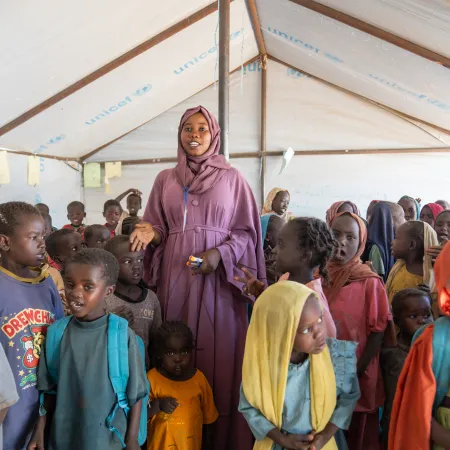 A teacher in a mauve robe stands among young children in a classroom within a UNICEF tent. Children wear colorful clothing.