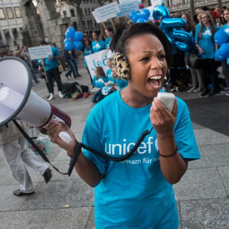 A girl wearing a blue UNCIEF tshirt speaks into a speakerphone, behind her others can be seen standing in a line also wearing UNICEF tshirts. 